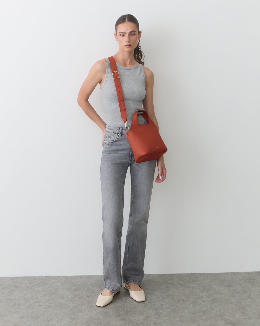 Person wearing tank top and jeans with shoulder bag standing against a plain wall.
