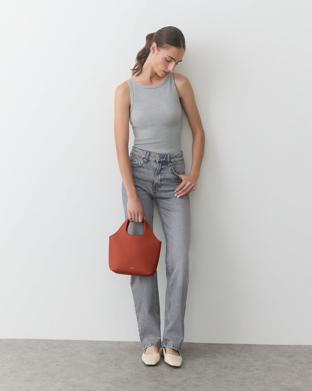 Person in tank top and jeans holding a small bag, standing against a plain background.