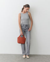 Person in tank top and jeans holding a small bag, standing against a plain background.