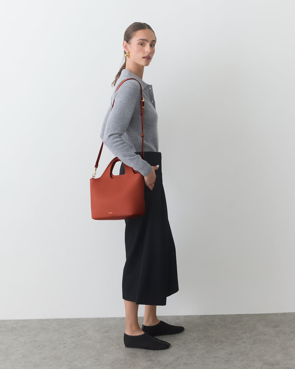 Person standing with a shoulder bag, wearing a long skirt and long-sleeved top.
