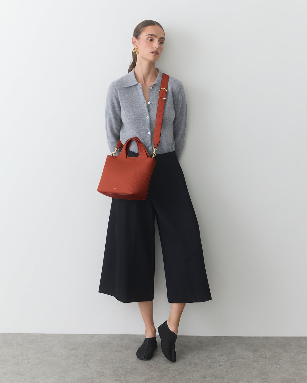 Person in long pants and shirt, carrying a shoulder bag, standing against light background.