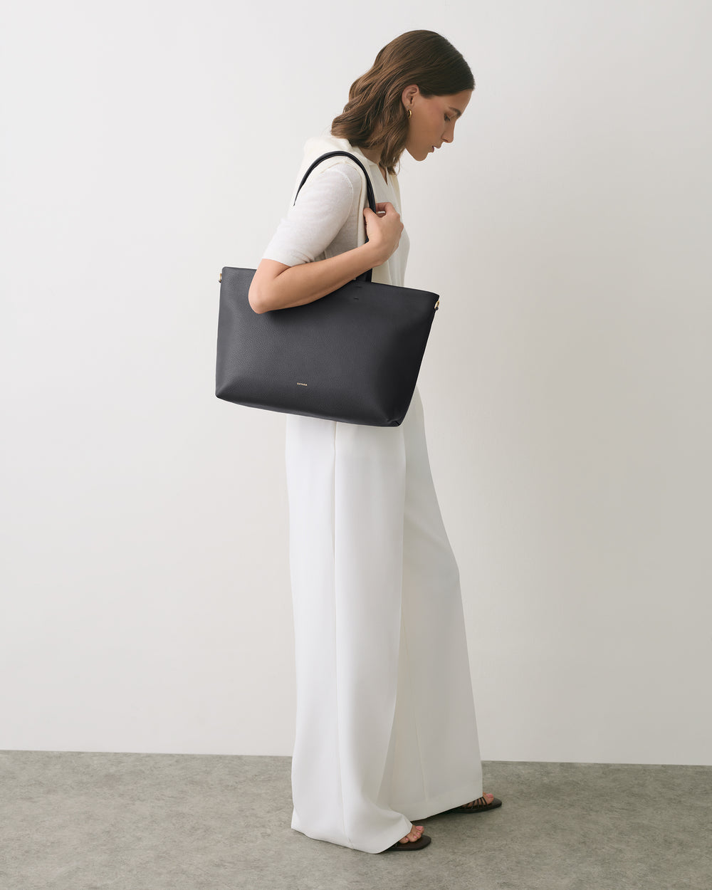 Person standing with tote bag over shoulder, wearing long pants and short-sleeved top.