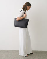 Person standing with tote bag over shoulder, wearing long pants and short-sleeved top.