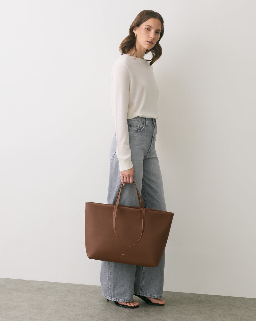 Person standing with tote bag, wearing a long-sleeve top and jeans.