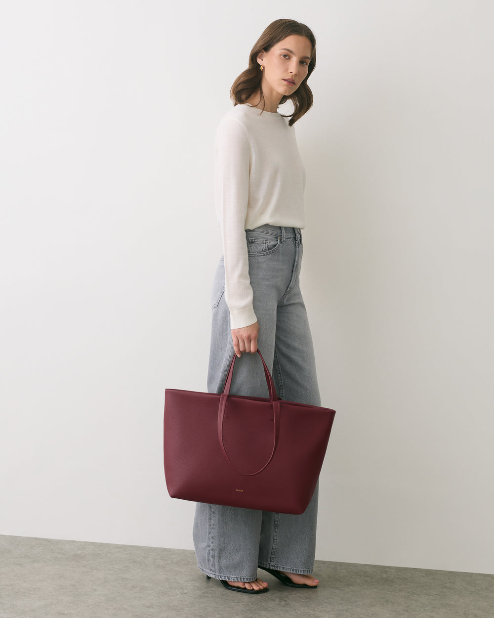 Person with a large handbag wearing a sweater and jeans, standing against a plain wall.