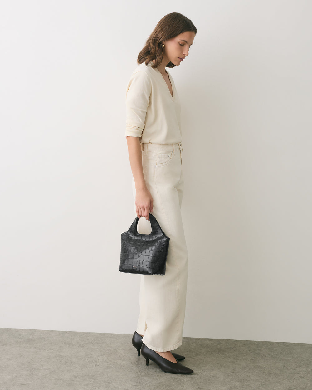 Person in outfit holding small croc-embossed leather handbag, wearing heels, standing sideways.