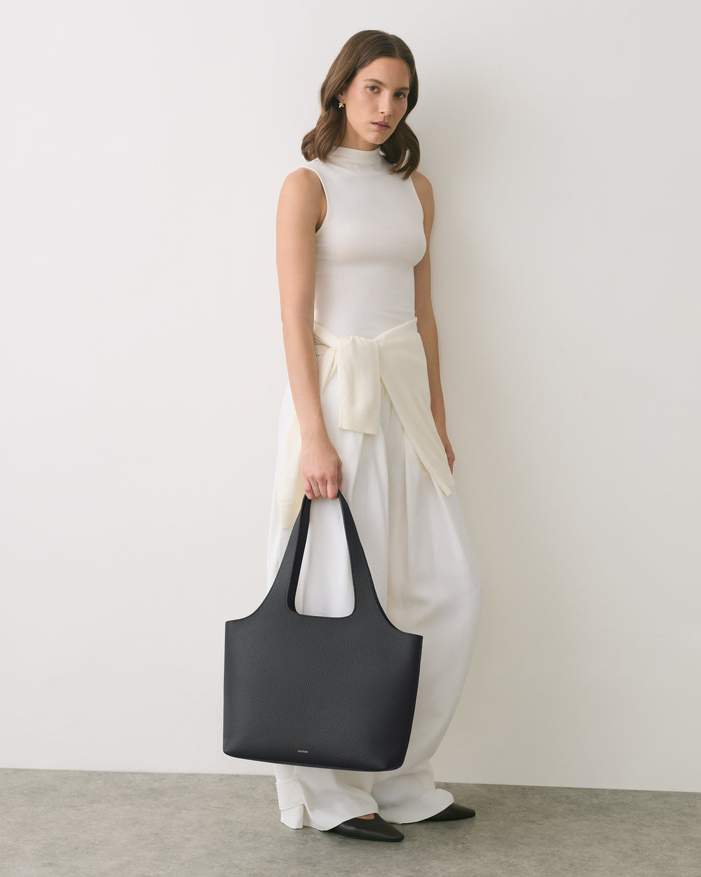 Person standing against a wall holding a large handbag, wearing a sleeveless top and long pants.