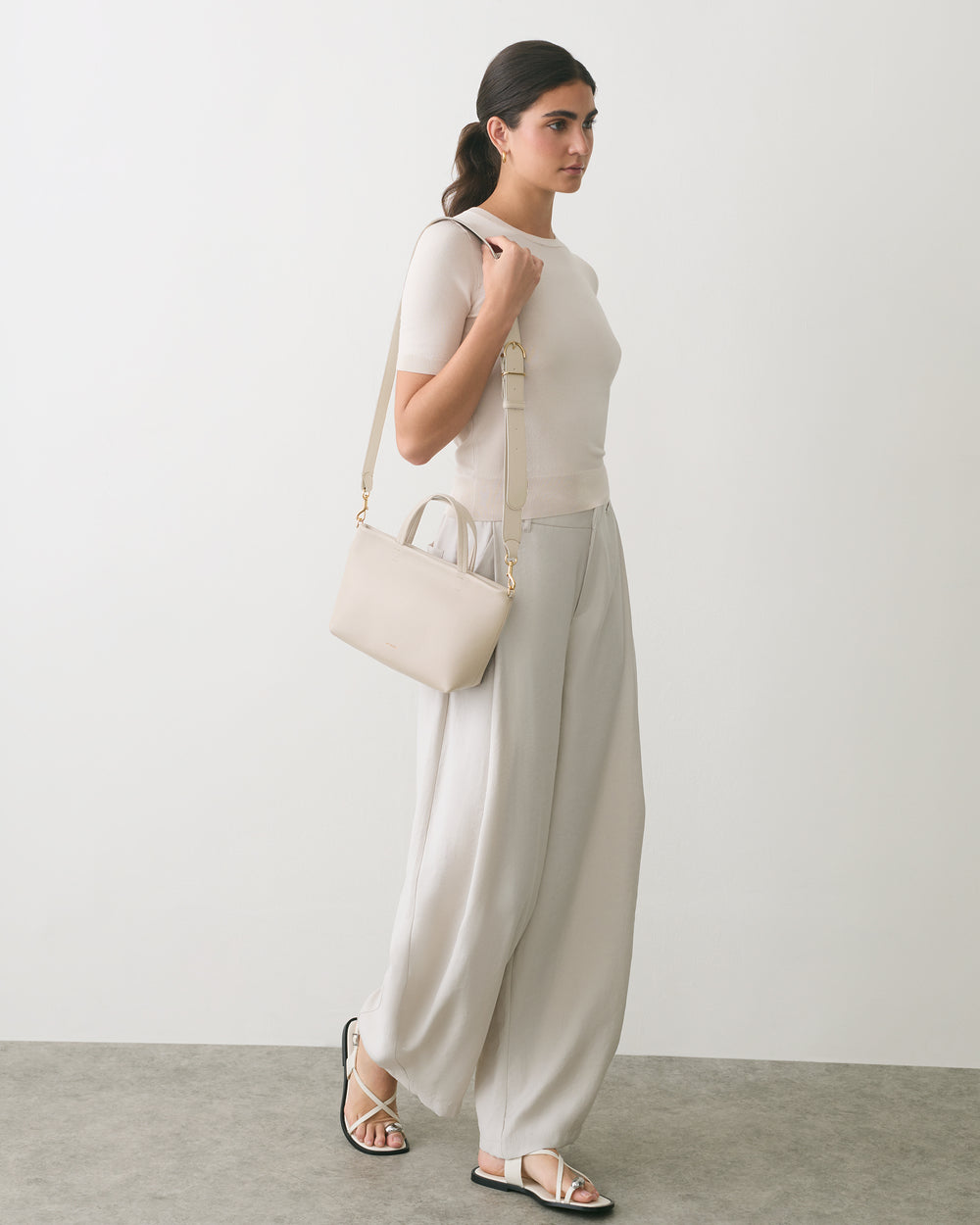 Person standing in profile, holding a shoulder bag, wearing loose-fitting pants and sandals.