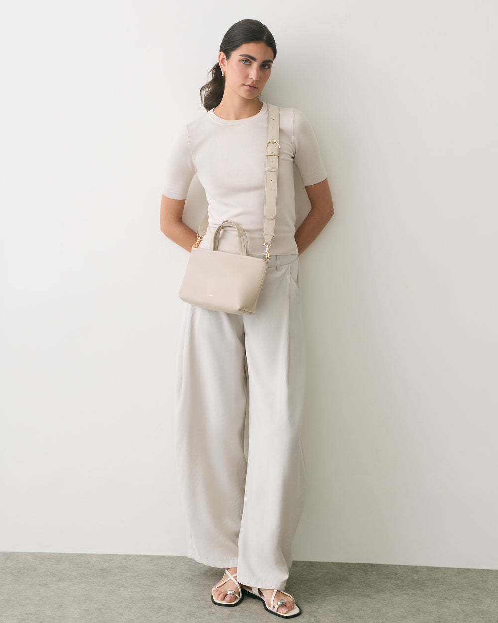 Person stands against wall, carrying a handbag, wearing a short-sleeve top and wide-leg pants.