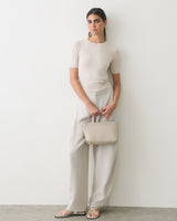 Person in a simple outfit holding a clutch bag, leaning against a plain wall.