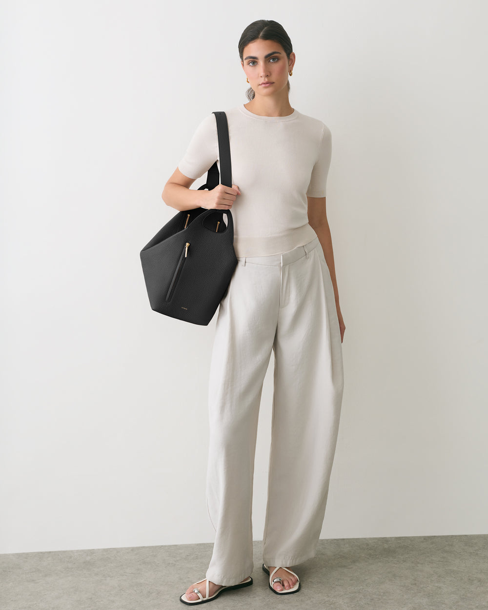 Person standing, holding a handbag, wearing a short-sleeve shirt and wide-leg pants, sandals.