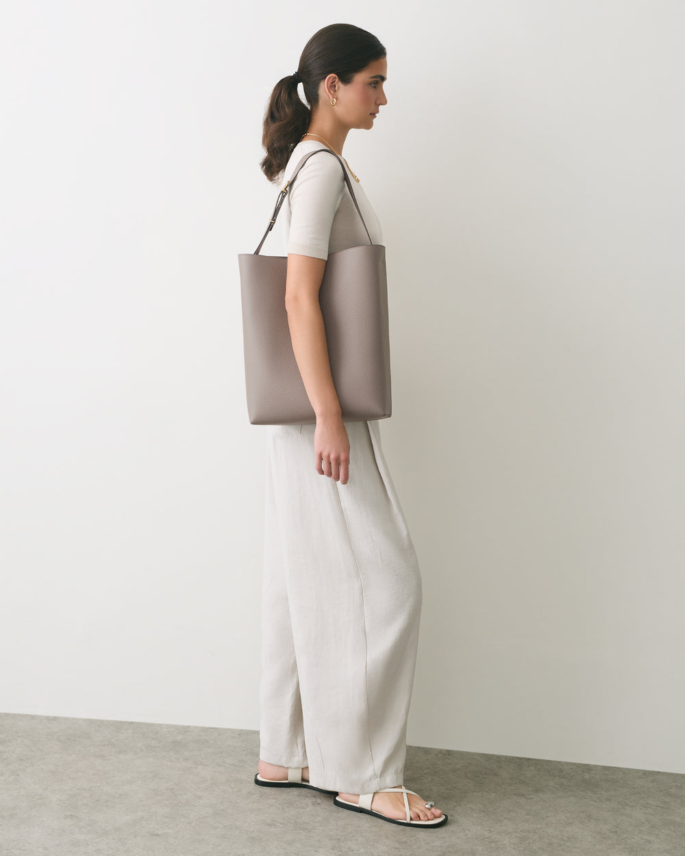 Woman standing sideways holding a shoulder bag wearing a casual outfit with sandals.