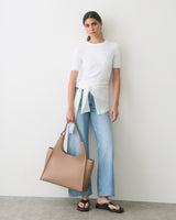 Person in jeans and tied shirt holding large tote bag, standing against plain wall.