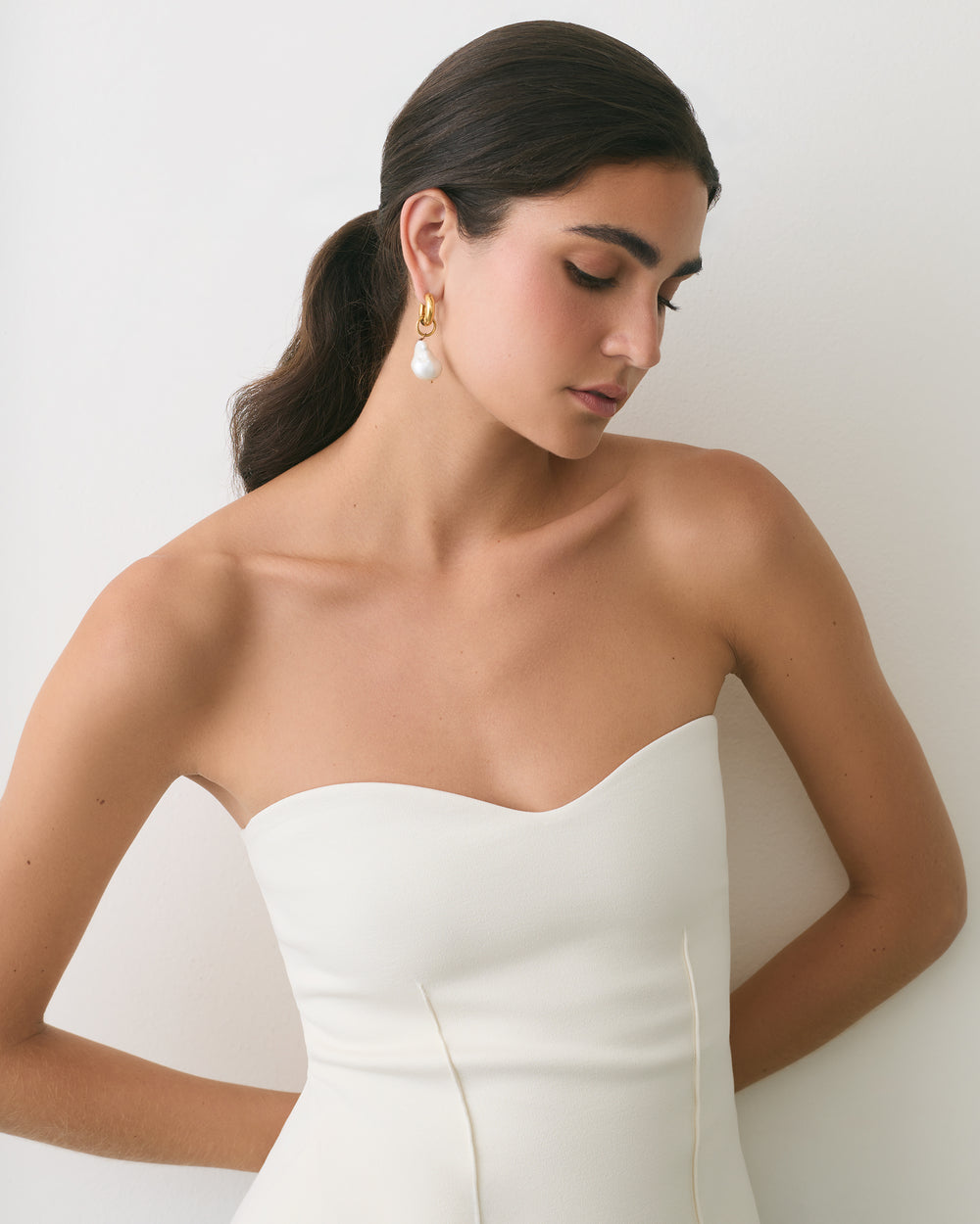 Person in strapless dress with earrings standing against a wall.