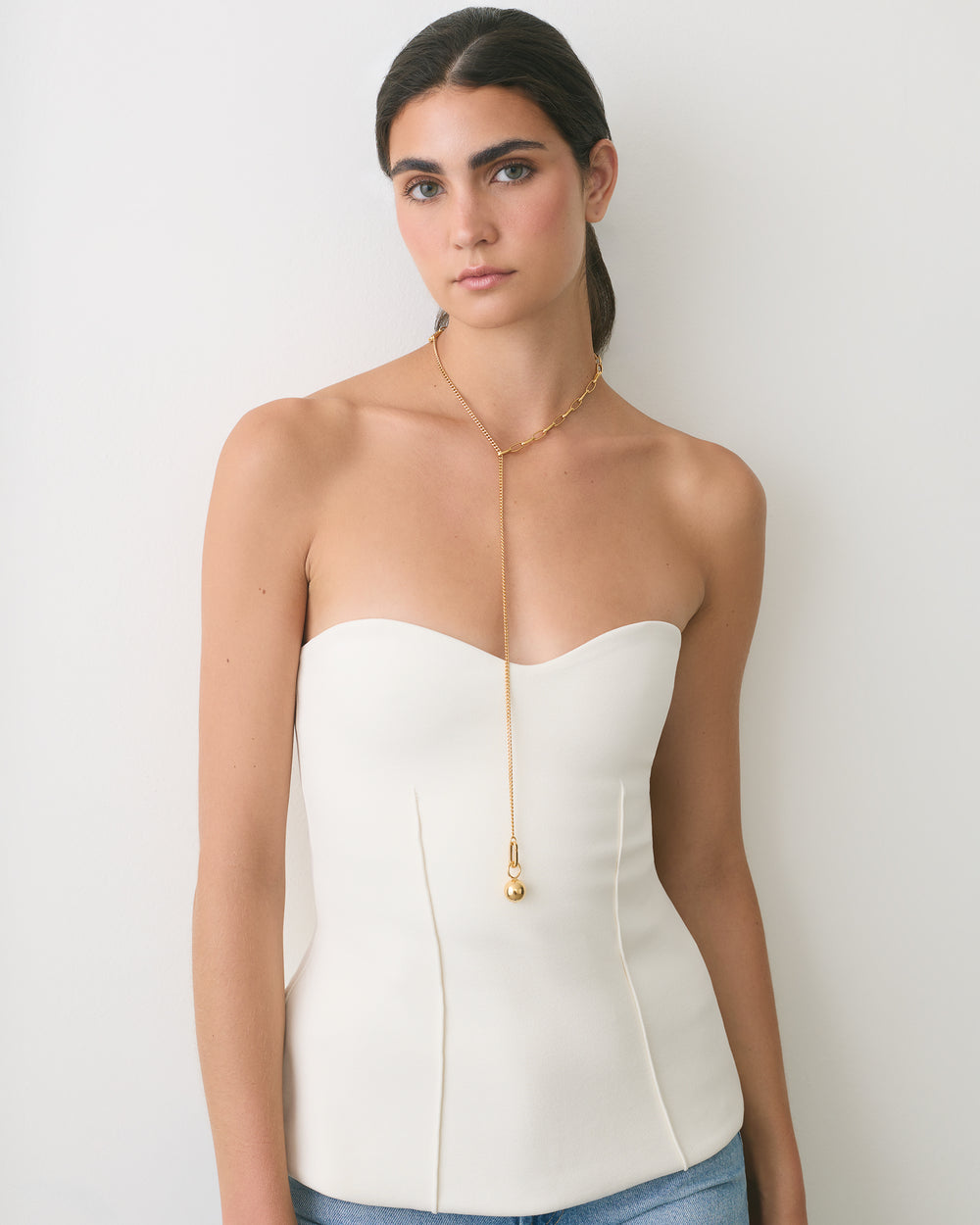Person wearing a strapless top with long necklace standing against a wall.