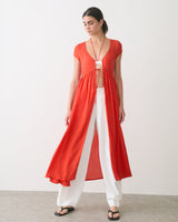 Person wearing a long open-front dress over a top and pants, standing indoors.