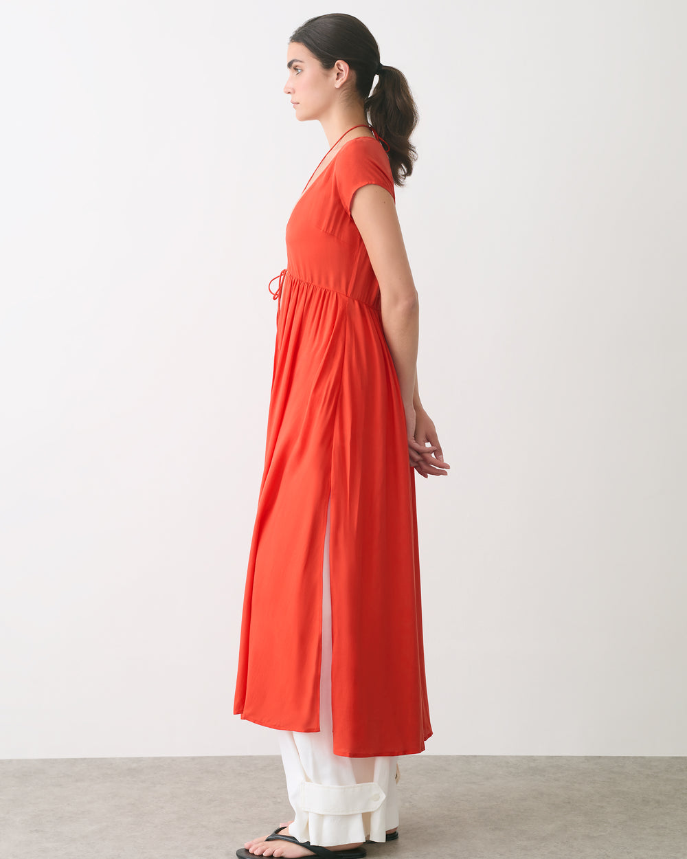 Woman in a long dress over pants, standing sideways in a neutral room.