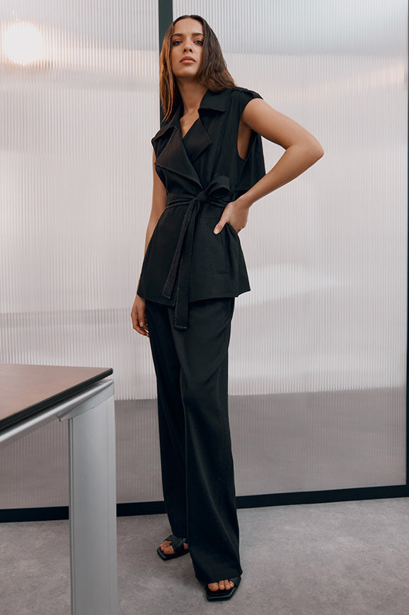 Model wearing a sleeveless belted top and wide-legged pants, standing indoors by a table.