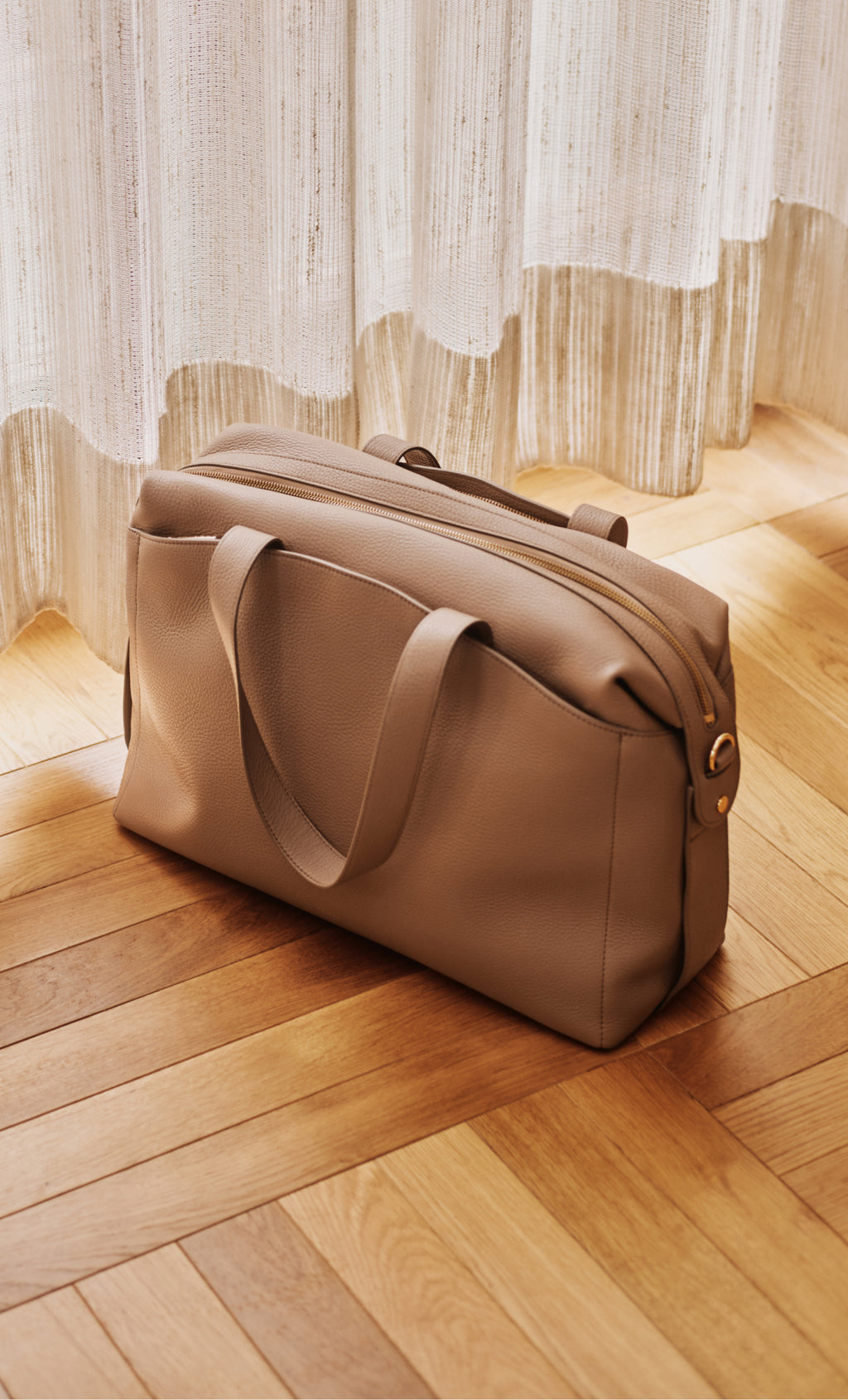 Travel bag on a wooden floor beside sheer curtains.