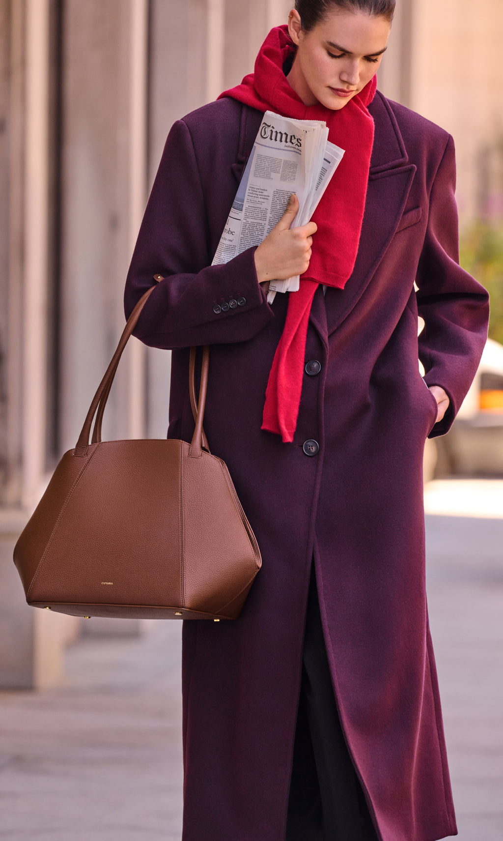 Person in long coat with scarf holding a newspaper and a large handbag walking outside