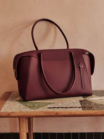 A structured burgundy leather handbag with double top handles and a vertical zip detail, displayed on a wooden table against a textured beige wall.