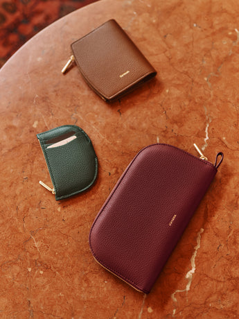 Three pebbled leather accessories arranged on a warm, textured terracotta marble surface: a brown zip-around wallet, a dark green card holder, and a deep burgundy zip-around pouch. Each item features a small gold-toned logo and a gold zipper pull.