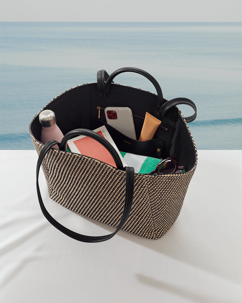 Open tote bag with various items inside on a white surface near a water body.