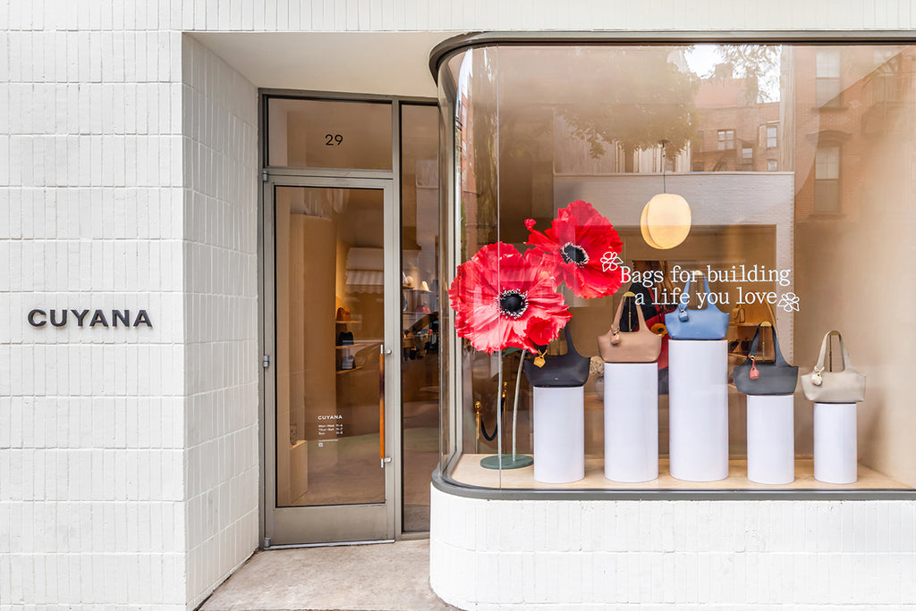 Storefront window displaying bags on pedestals with a text sign and red flowers.