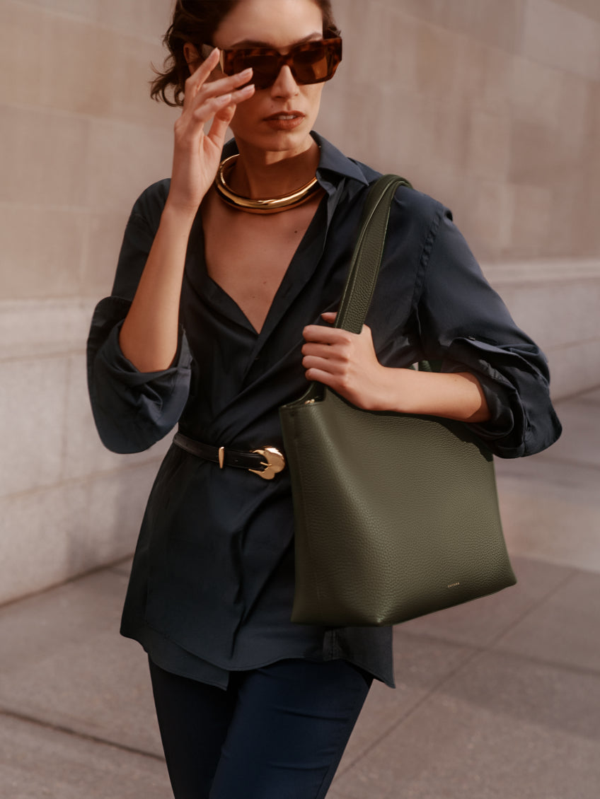 Person walking, wearing oversized sunglasses, holding a shoulder bag, and touching their face.