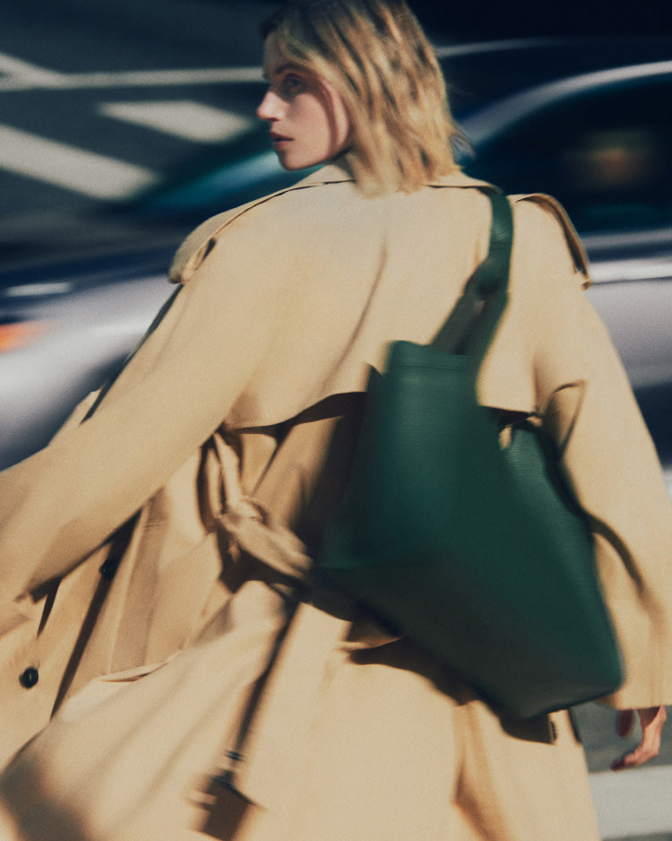 Person walking in a city wearing a long coat and carrying a large bag.