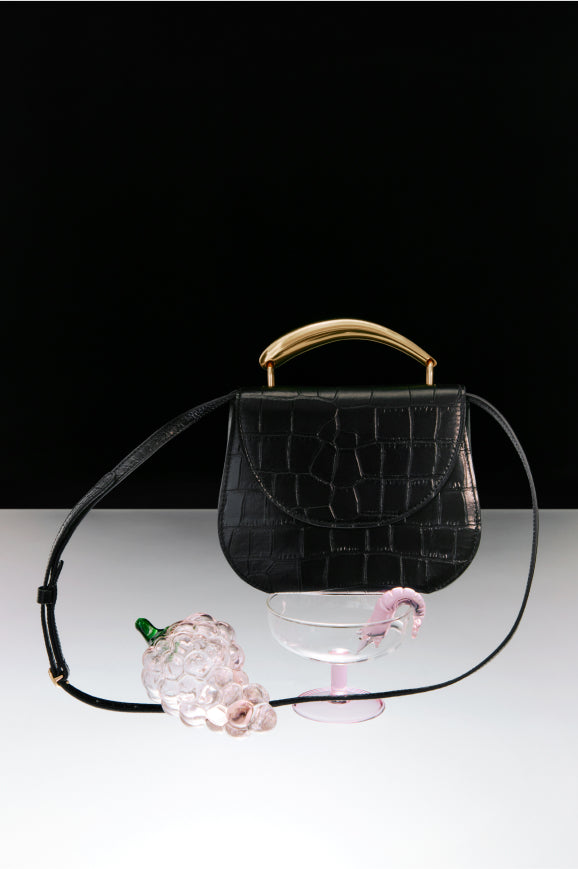 Handbag with a shoulder strap, a perfume bottle, and a glass on a table.