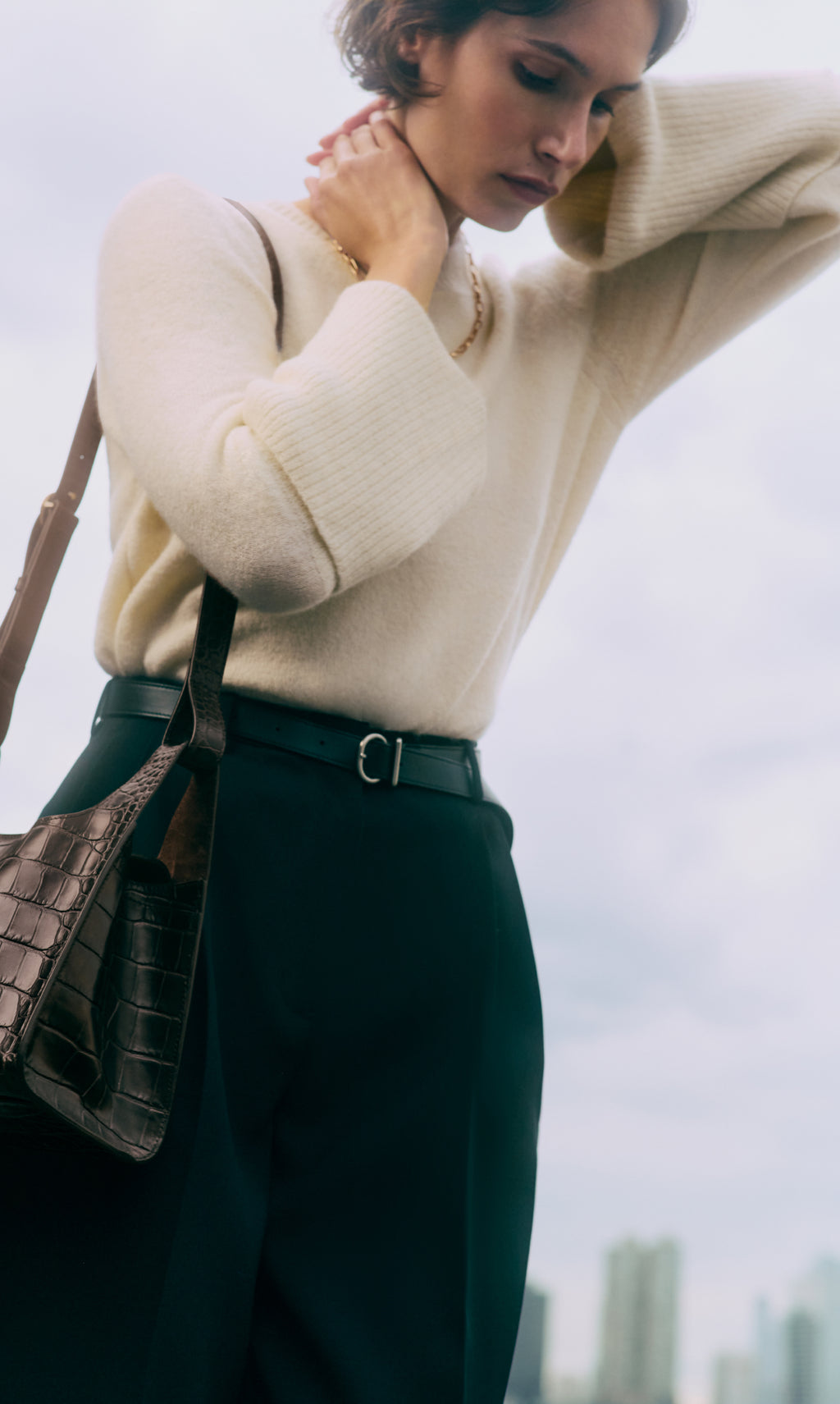 Person with sweater and pants holds croc-embossed leather bag, cloudy sky in background.