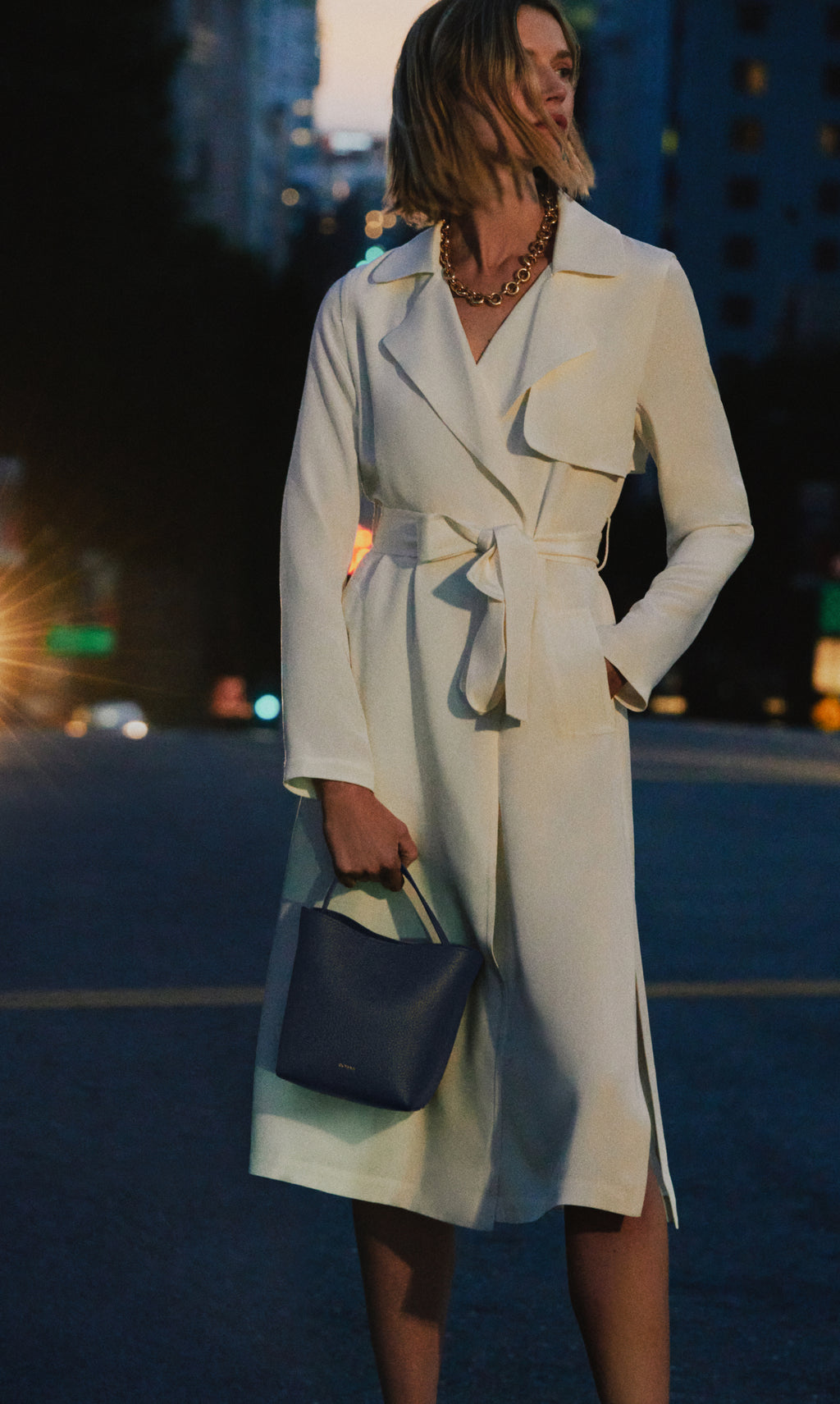 Person in a trench coat holds a handbag, standing on a city street at dusk.