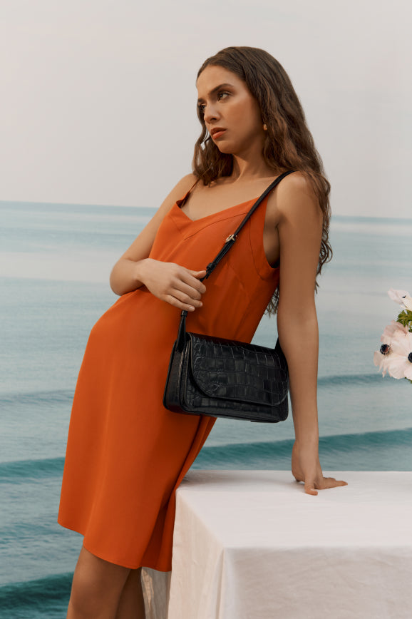 Woman standing by a table with a purse, looking away, ocean in the background.