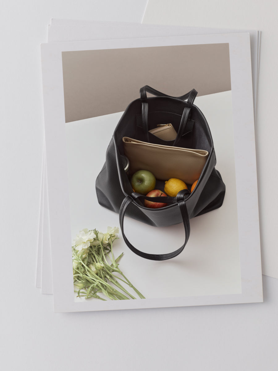 Open handbag with a wallet, apples, oranges, and flowers on a white surface.