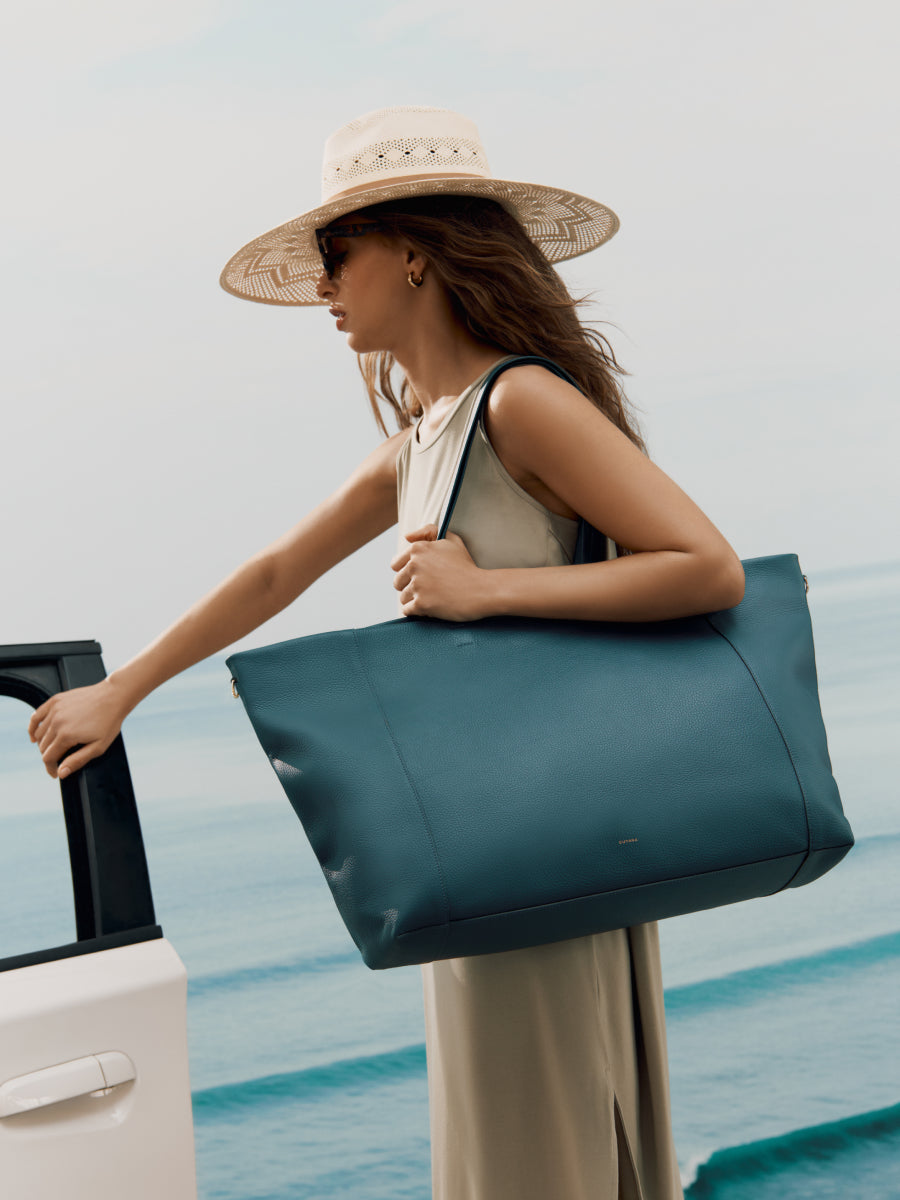 Person in a dress and hat holding a large bag next to a car by the seaside