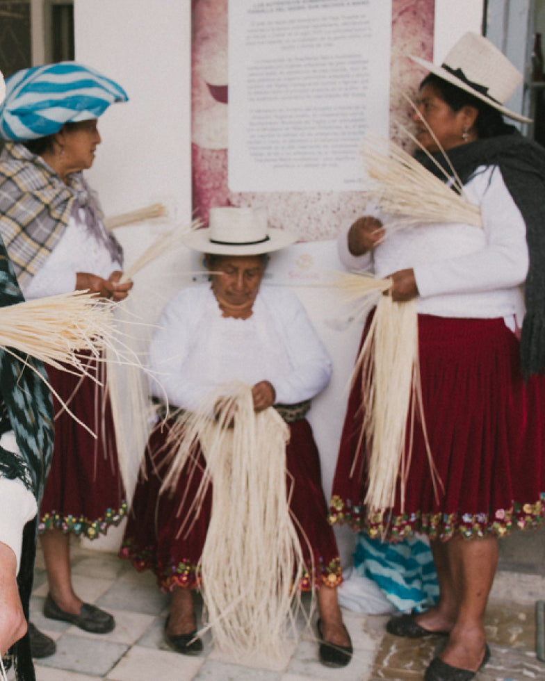 People weaving straw items while standing and sitting near an informational plaque.
