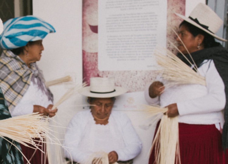 Three people weaving straw hats in front of a wall with text.