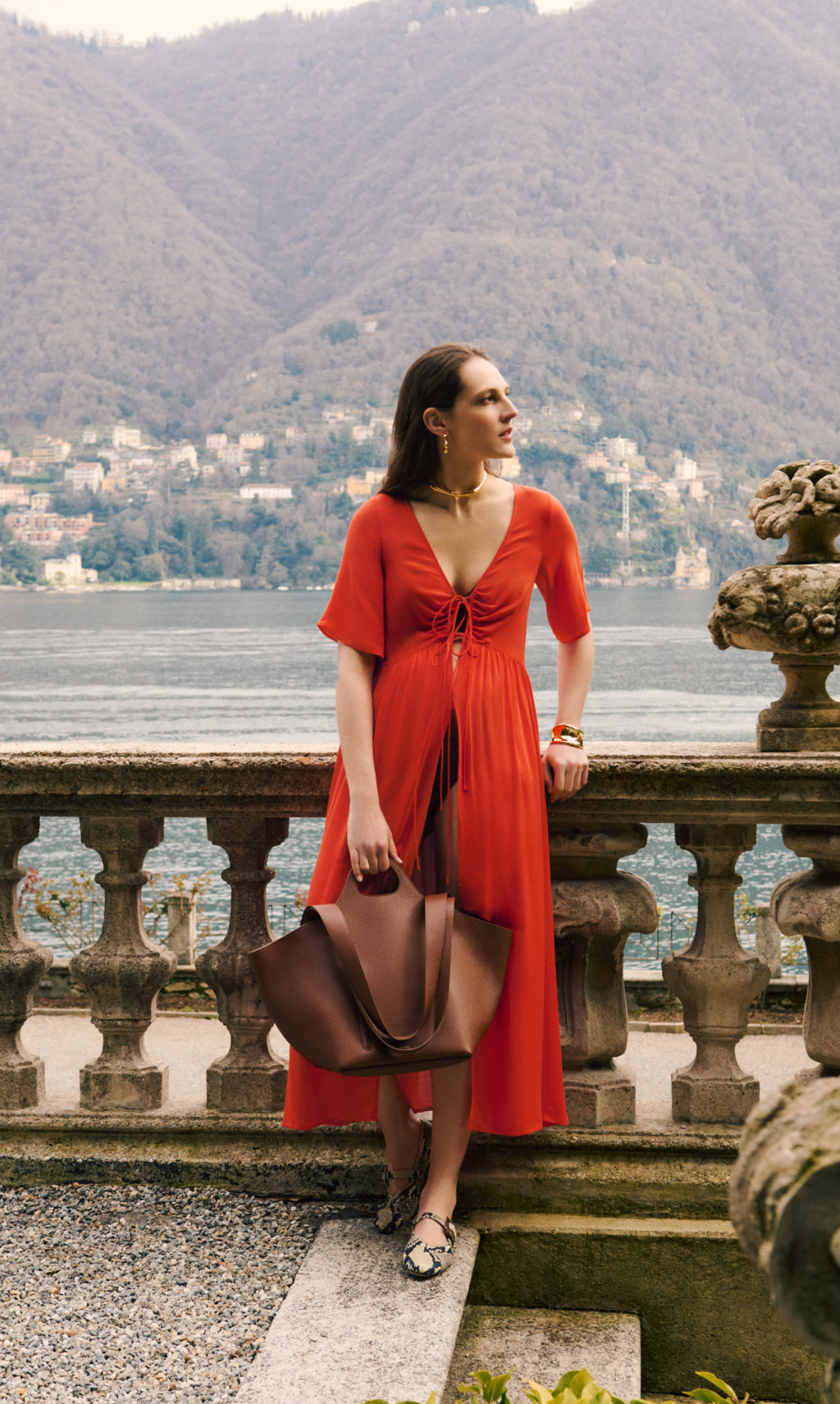 Woman in a dress holding a large tote bag, standing by a railing with a lake and mountains in the background.