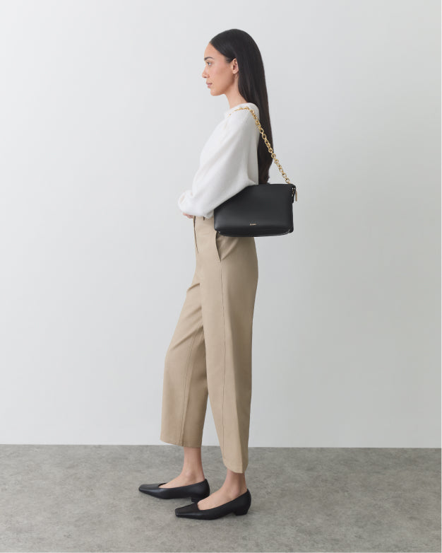 Person standing sideways with a shoulder bag and wearing pants and long-sleeved top.