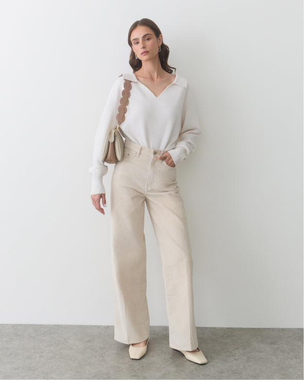 Woman in a long-sleeve top and wide-leg pants holding a purse, standing against a wall.
