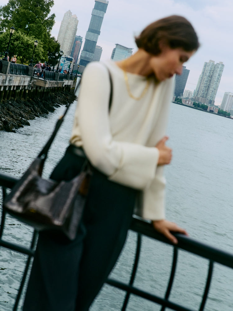 Person leaning on a railing by the water with a croc-embossed leather shoulder bag.