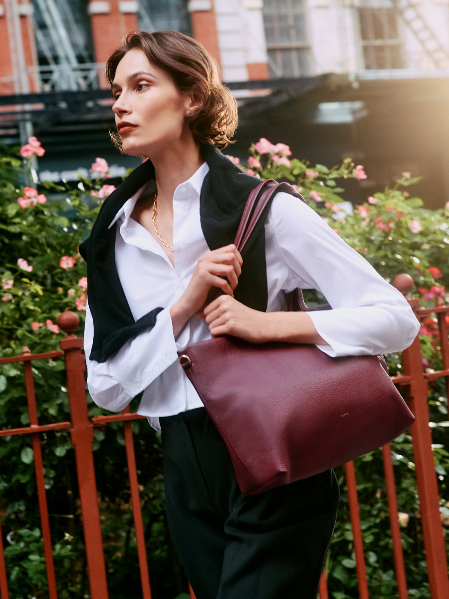 Person holding a shoulder bag, wearing a long sleeve shirt and sweater over shoulders, with flowers in background