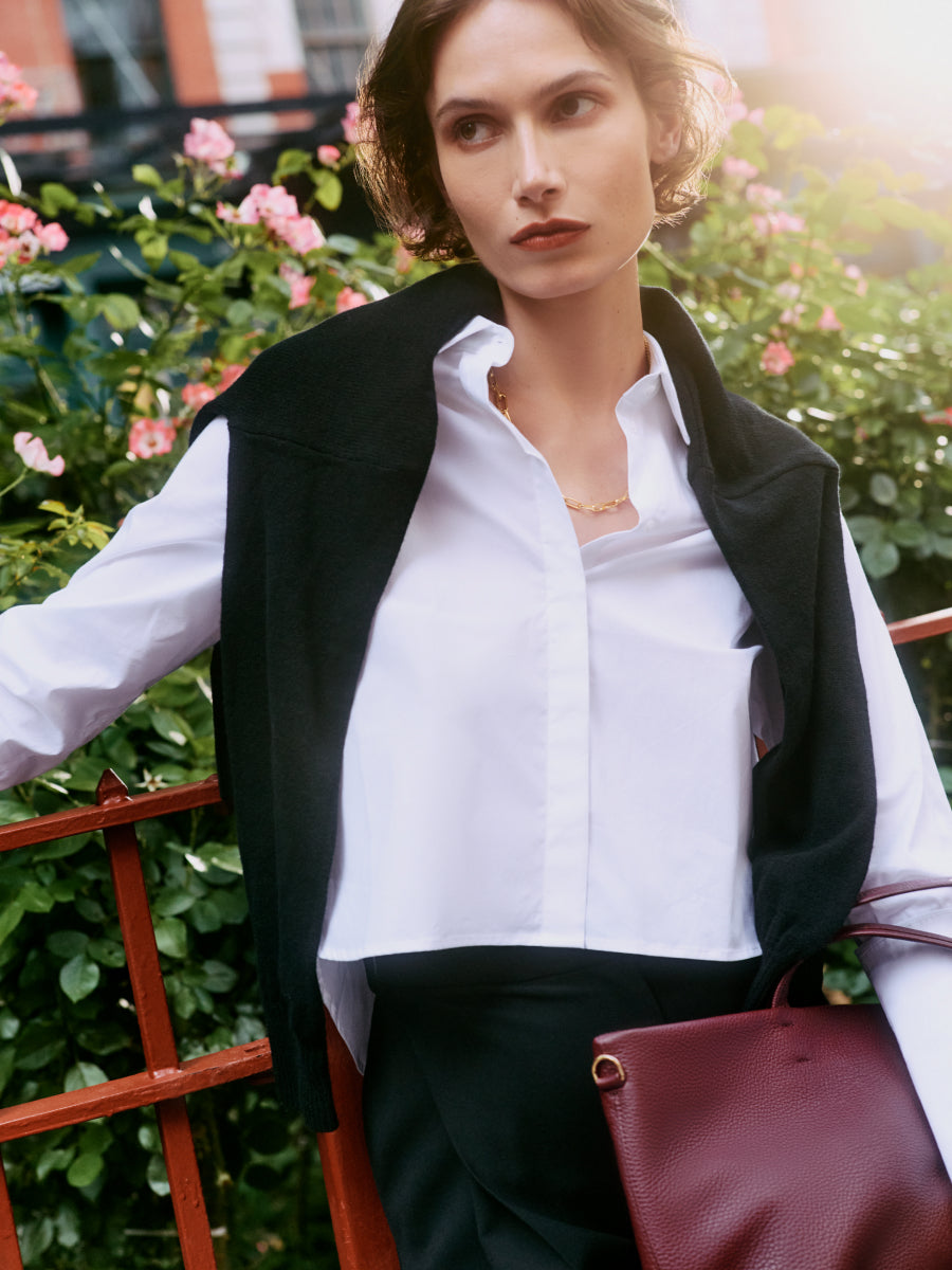 Person in a white shirt and black jacket over shoulders, holding a bag, standing by a red fence.