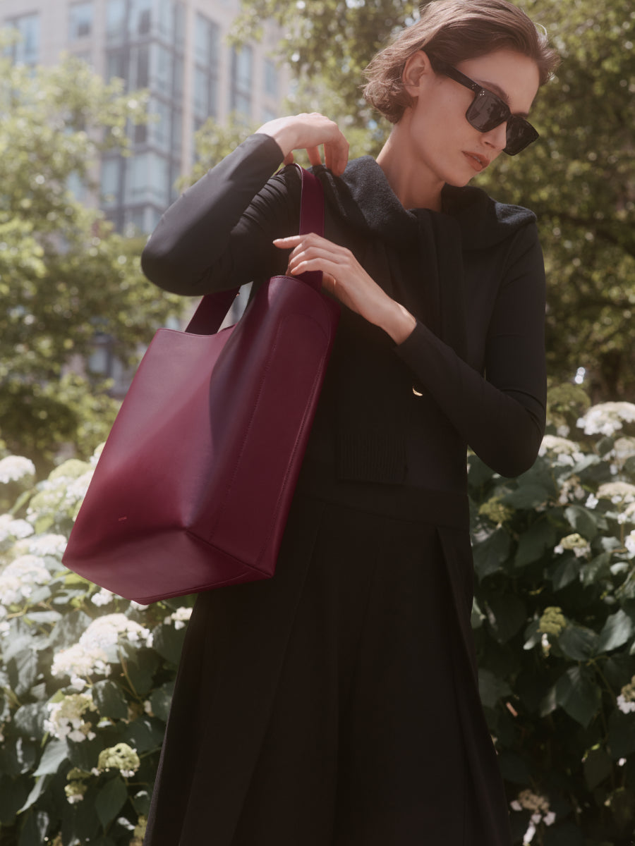 Person holding a tote bag in an outdoor setting with plants and buildings in the background.