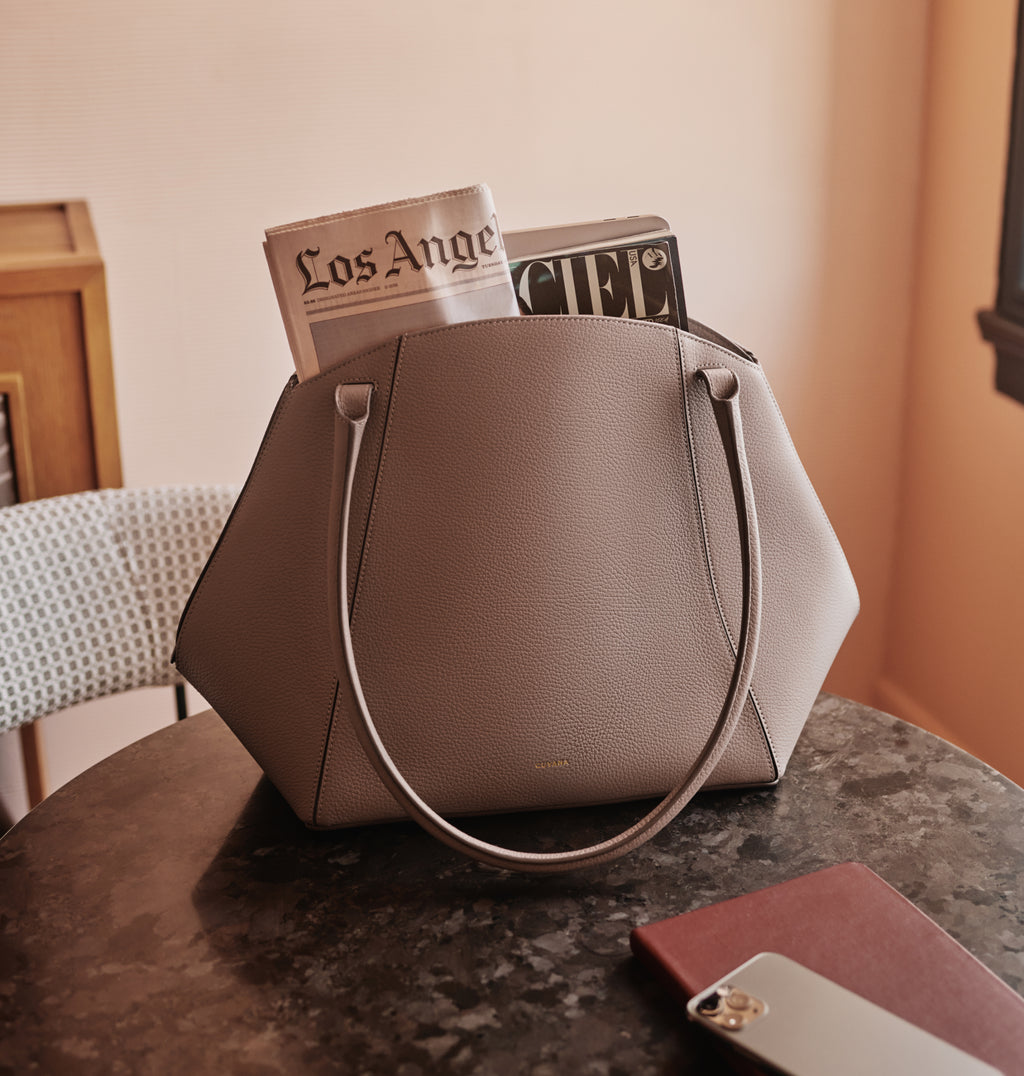 Handbag on table with magazines inside, next to a phone and closed notebook.