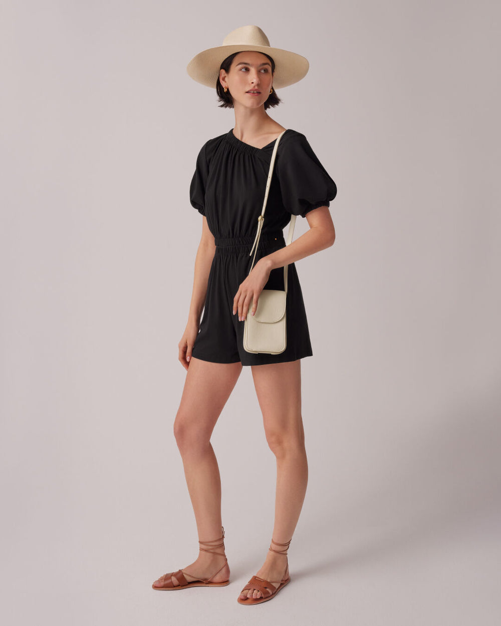 Woman standing in a romper with a hat, holding a shoulder bag and wearing sandals.