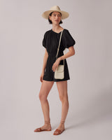 Woman standing in a romper with a hat, holding a shoulder bag and wearing sandals.