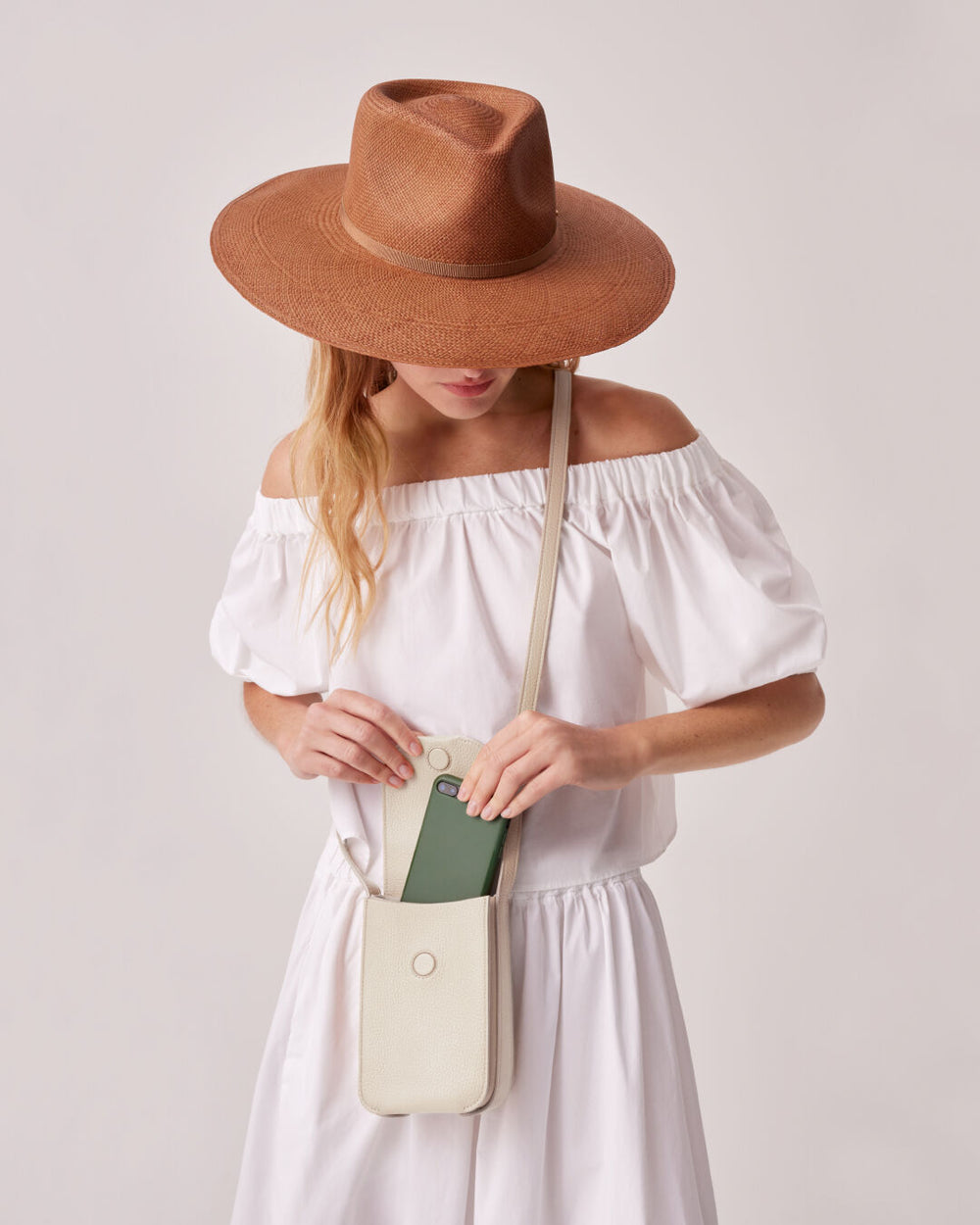 Woman in a wide-brimmed hat and dress putting a phone in a shoulder bag.