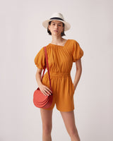 Woman in a romper and hat standing with one hand on hip and holding a round bag.